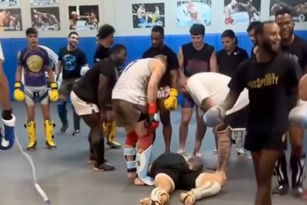 Lutadores cercam um homem nocauteado no chão da academia durante uma sessão de sparring.