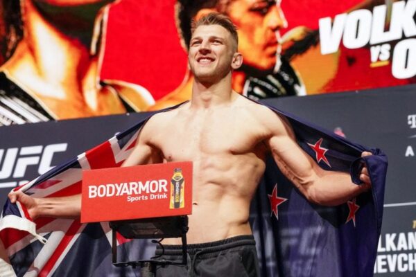 Lutador do UFC Dan Hooker sorri na pesagem com a bandeira da Nova Zelândia sobre os ombros.