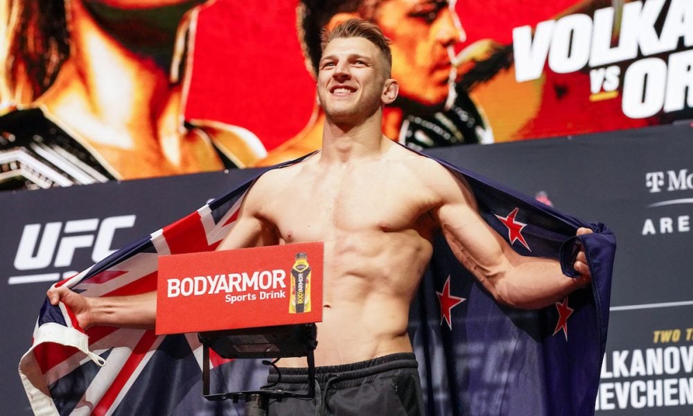 Lutador do UFC Dan Hooker sorri na pesagem com a bandeira da Nova Zelândia sobre os ombros.