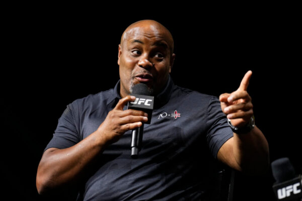 UFC Hall of Famer Daniel Cormier speaks into a microphone, gesturing with his finger against a dark background.