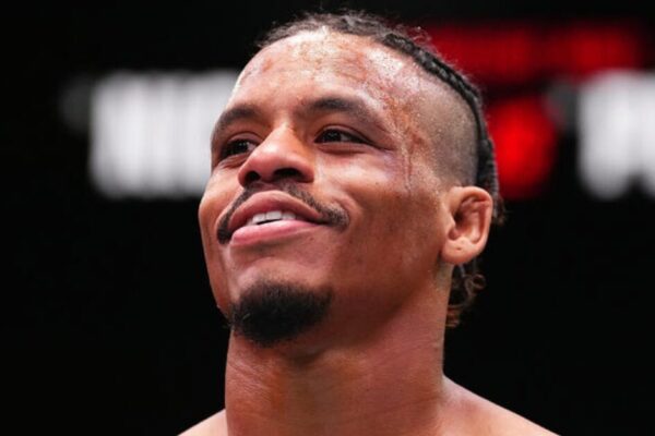 A smiling Black MMA fighter with braided hair and a sweaty forehead looks up.
