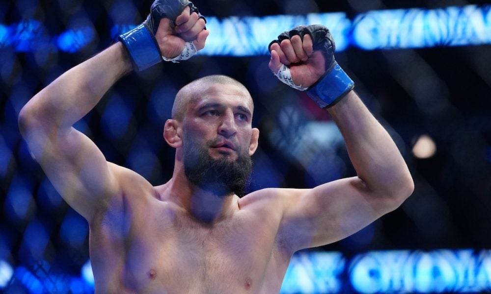 A shirtless Khamzat Chimaev with a beard raises his gloved fists in victory inside the UFC octagon.