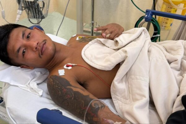 A man with tattoos on his arm and chest lies in a hospital bed with medical sensors attached, looking at the camera.