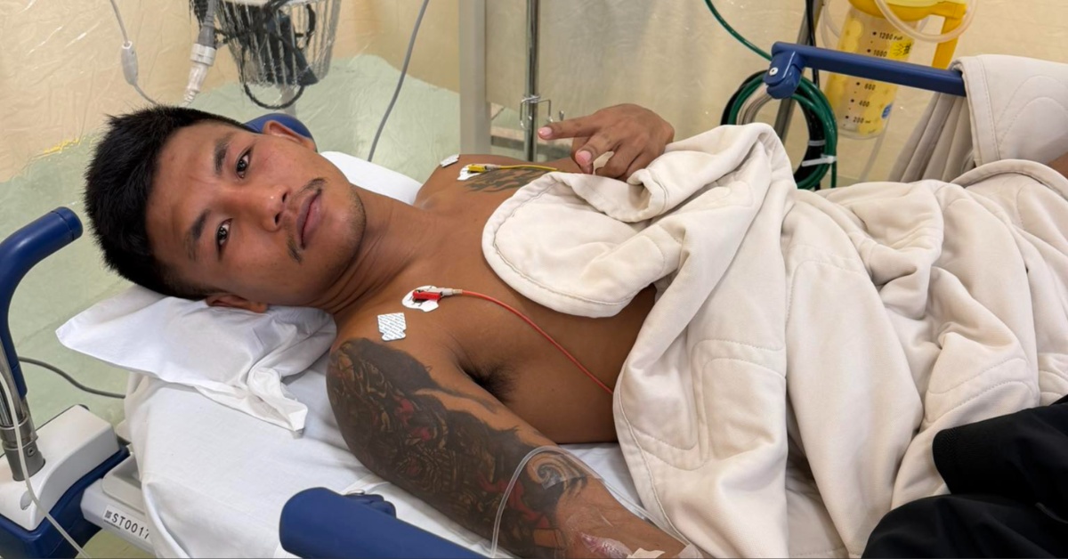 A man with tattoos on his arm and chest lies in a hospital bed with medical sensors attached, looking at the camera.