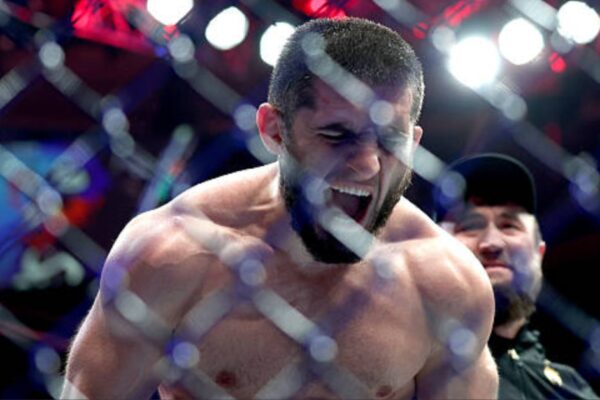 MMA fighter Islam Makhachev roars intensely inside the octagon, seen through the cage fence.