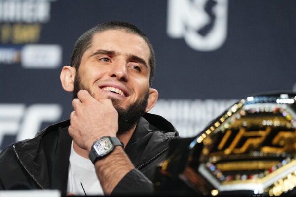 O campeão do UFC, Islam Makhachev, sorri em coletiva de imprensa com seu cinturão em primeiro plano.