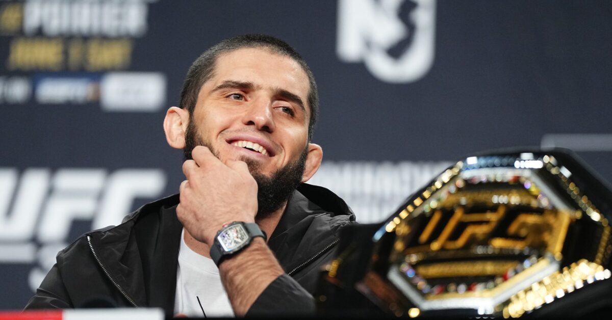 O campeão do UFC, Islam Makhachev, sorri em coletiva de imprensa com seu cinturão em primeiro plano.