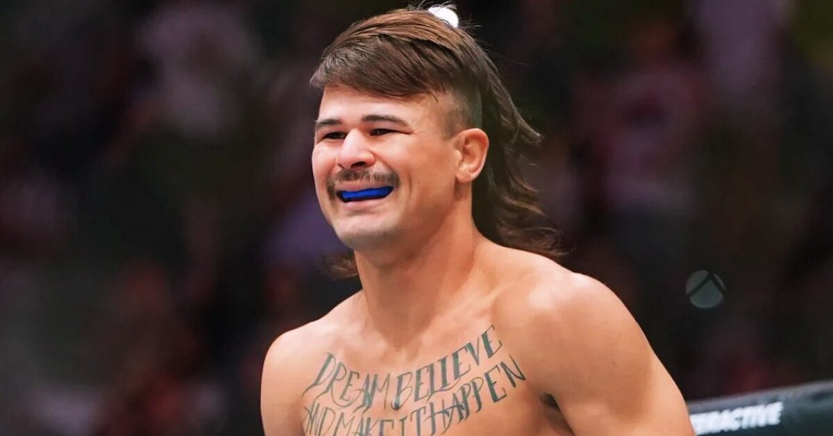 Close-up do lutador Myles Jury com bigode e mullet, sorrindo com um protetor bucal azul e tatuagem no peito.