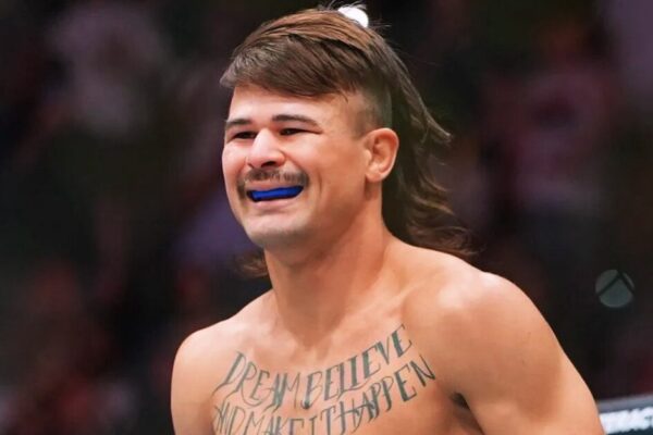 Close de um lutador com cabelo mullet e bigode, sorrindo com protetor bucal azul e tatuagem no peito.