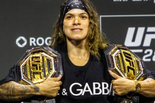 UFC double-champion Amanda Nunes smiles, holding two championship belts over her shoulders at a press event.
