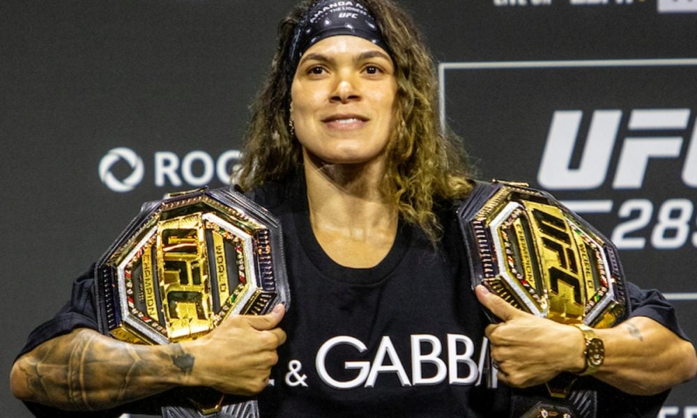 UFC double-champion Amanda Nunes smiles, holding two championship belts over her shoulders at a press event.