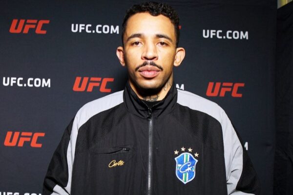 UFC fighter Carlos Prates in a black and grey jacket poses against a UFC step-and-repeat backdrop.