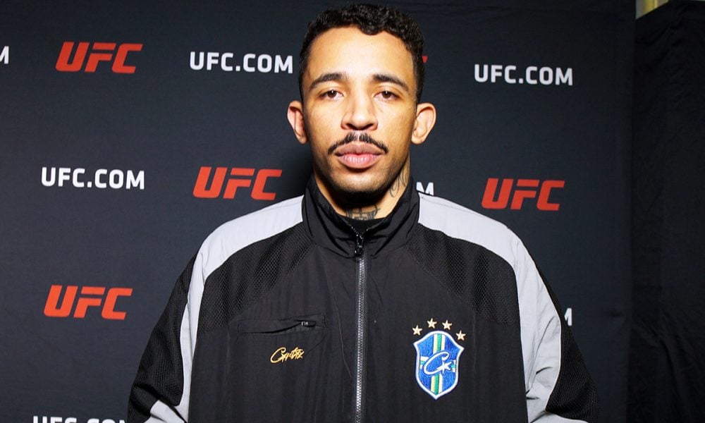 UFC fighter Carlos Prates in a black and grey jacket poses against a UFC step-and-repeat backdrop.