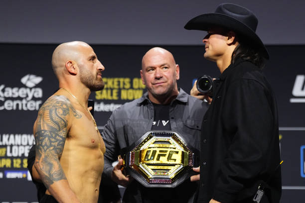 Dois lutadores do UFC se encaram com Dana White ao centro, segurando o cinturão de campeão.