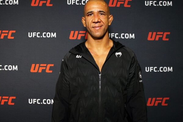 O lutador do UFC Gregory 'Robocop' Rodrigues sorri, vestindo uma jaqueta preta da Venum em frente a um banner do evento.