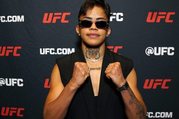 A lutadora do UFC Iasmin Lucindo posa para foto com óculos escuros e punhos cerrados em frente a um painel do evento.