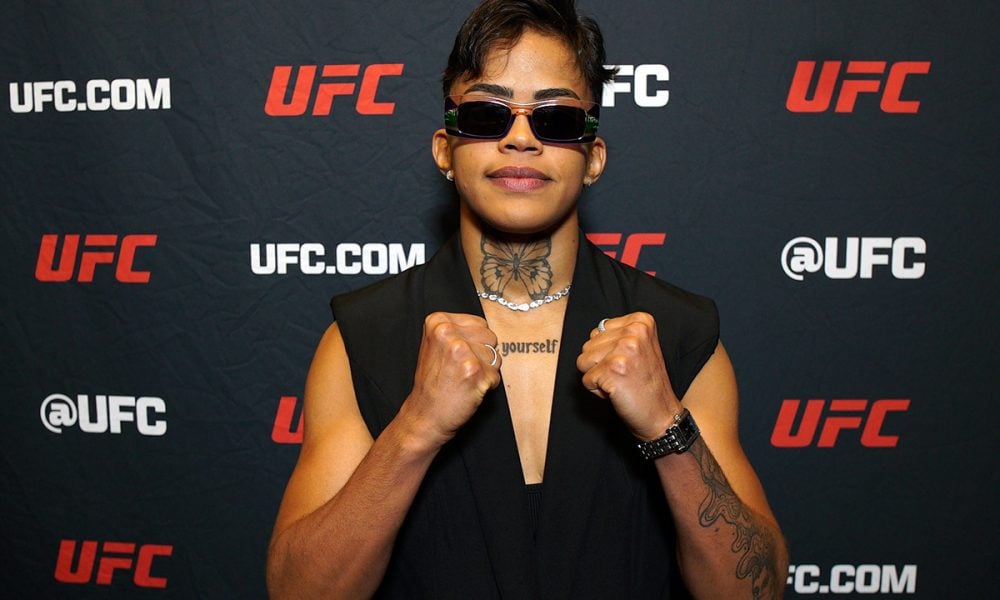 A lutadora do UFC Iasmin Lucindo posa para foto com óculos escuros e punhos cerrados em frente a um painel do evento.