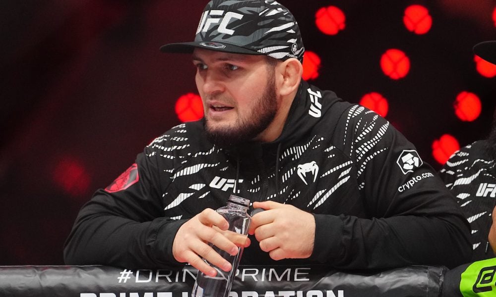 Khabib Nurmagomedov in a UFC cap and jacket coaches from the corner, holding a water bottle with a focused expression.