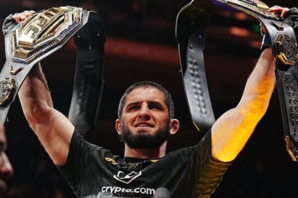 UFC champion Islam Makhachev smiles, holding two championship belts triumphantly above his head.