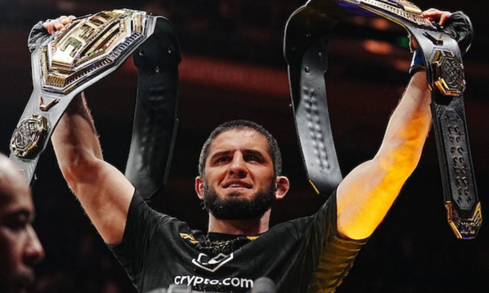 UFC champion Islam Makhachev smiles, holding two championship belts triumphantly above his head.