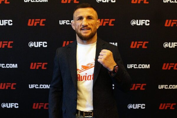 O lutador do UFC Merab Dvalishvili sorri e posa com o punho cerrado em frente a um painel com logos do UFC.