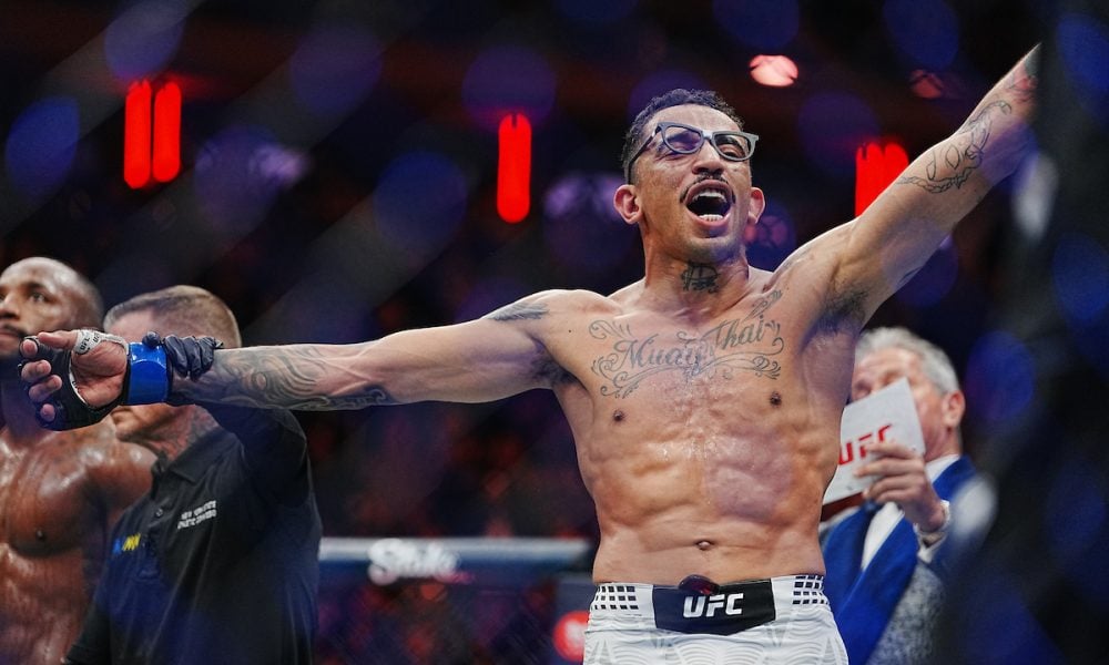Lutador do UFC de óculos e tatuagens comemora vitória com os braços abertos no octógono.