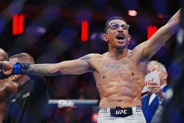 A tattooed UFC fighter with glasses roars in celebration, arms outstretched in the octagon after a victory.