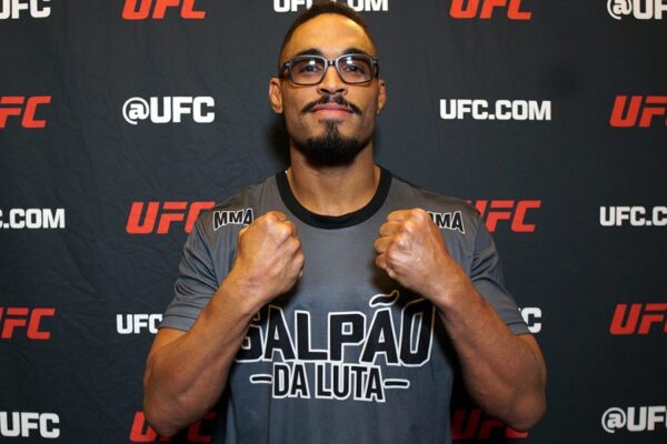 Lutador do UFC Rafeel Cerqueira, de óculos, posa com os punhos cerrados em frente a um painel do evento.