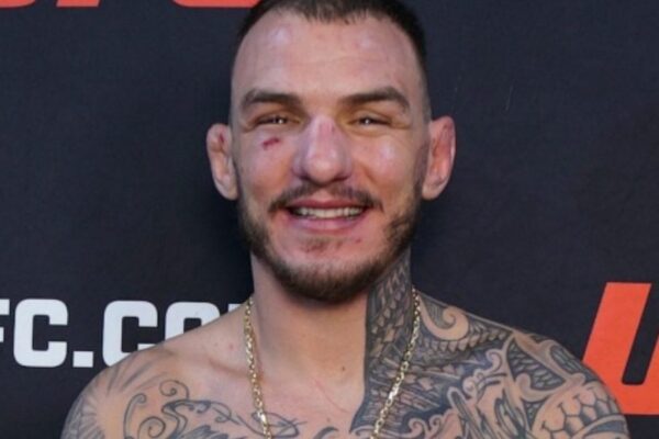UFC fighter Renato Moicano smiles in a headshot, shirtless with tattoos and a gold chain against a UFC backdrop.