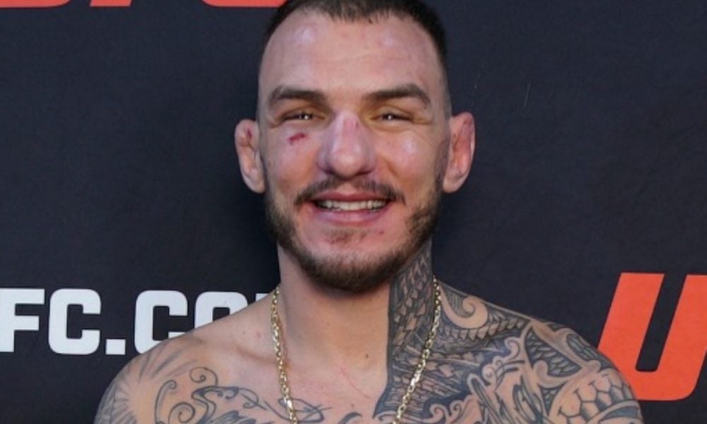 UFC fighter Renato Moicano smiles in a headshot, shirtless with tattoos and a gold chain against a UFC backdrop.