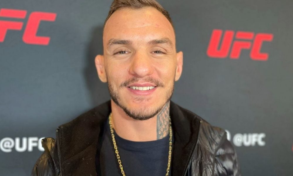 O lutador do UFC Renato Moicano sorri para a câmera, vestindo jaqueta preta em frente a um painel do evento.