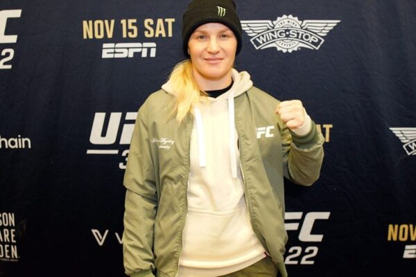 UFC fighter Valentina Shevchenko in a green jacket and beanie poses with her fist up in front of a UFC event backdrop.