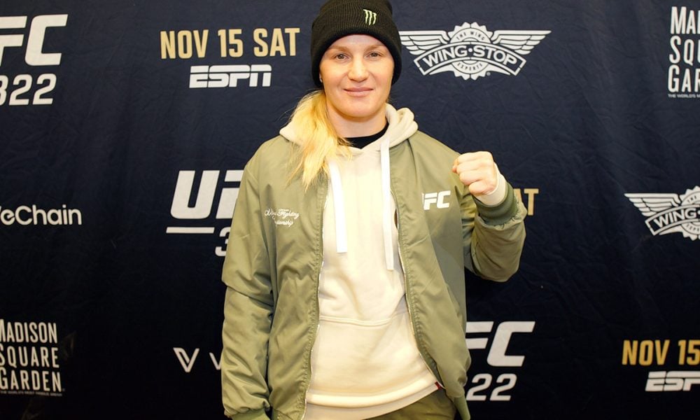 UFC fighter Valentina Shevchenko in a green jacket and beanie poses with her fist up in front of a UFC event backdrop.