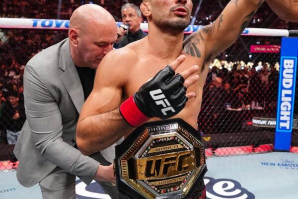 Alexandre Pantoja, campeão do UFC, comemora com o cinturão e o braço erguido no octógono.