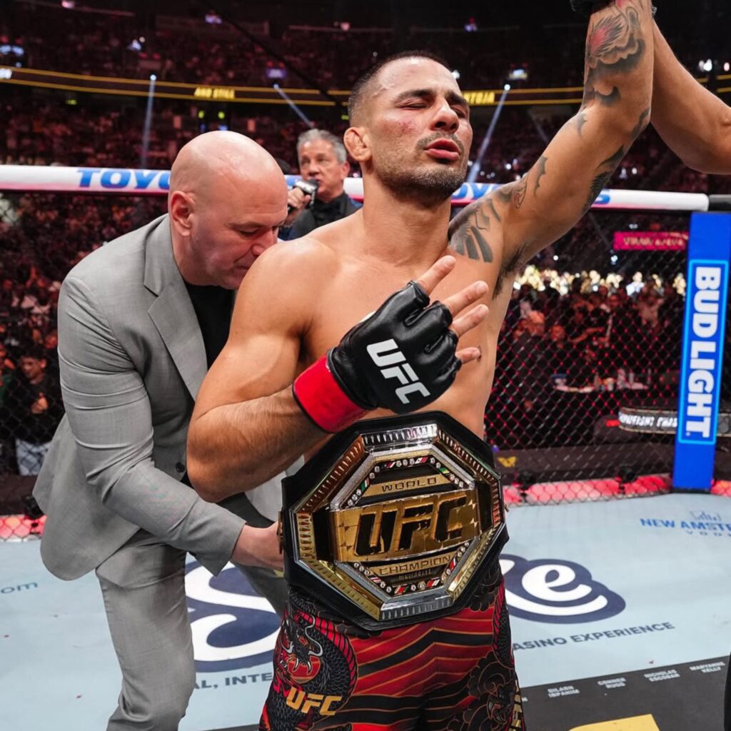 Alexandre Pantoja, campeão do UFC, comemora com o cinturão e o braço erguido no octógono.