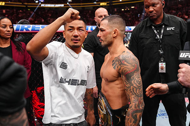 Lutador com punho erguido encara o campeão do UFC com cinturão e tatuagens, dentro do octógono.